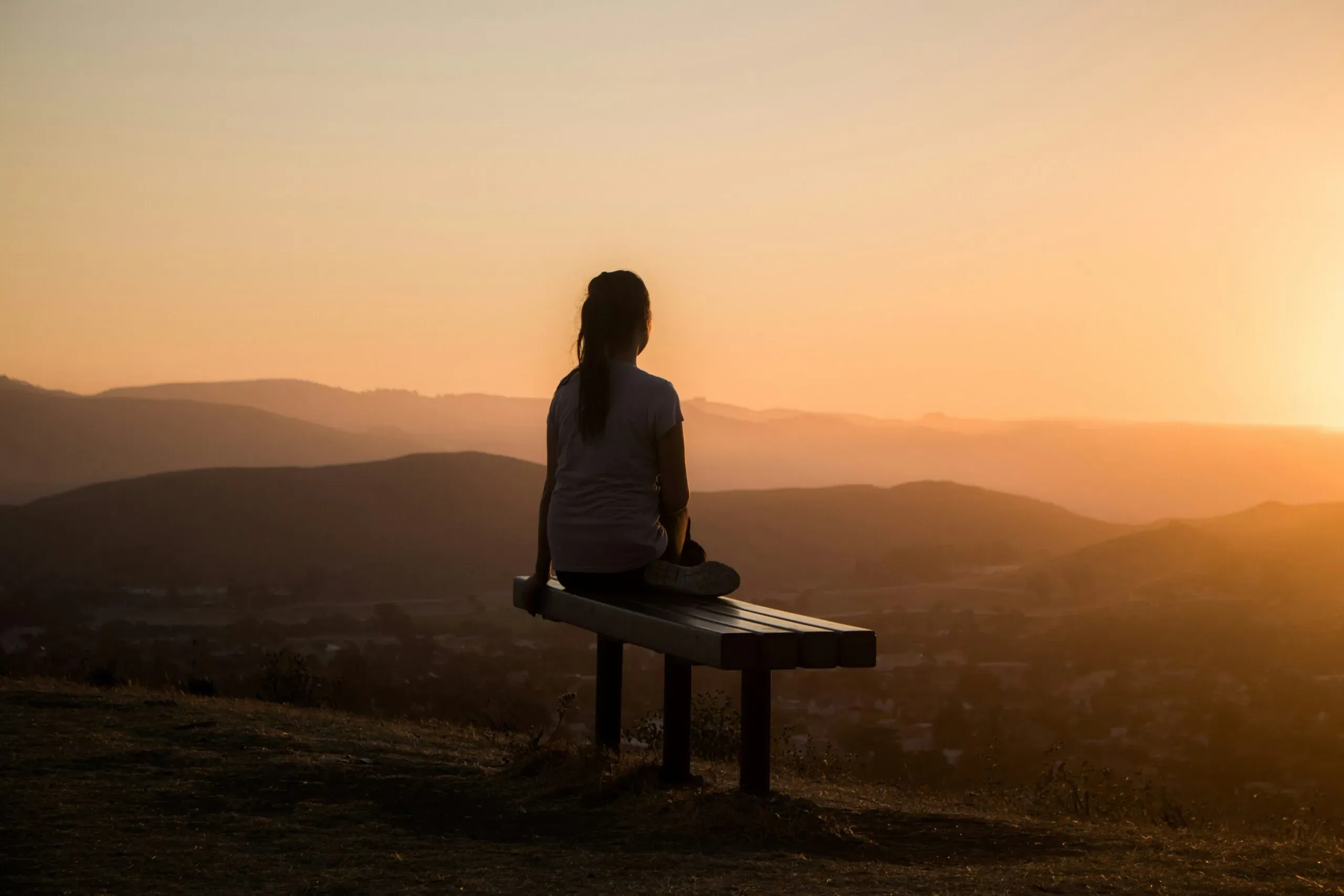 Silhouette d'une personne assise sur un banc face au coucher de soleil sur les collines.